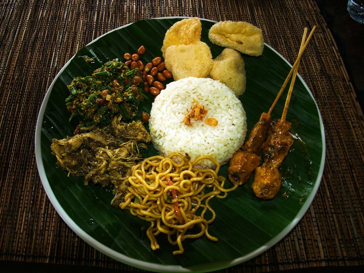 A plate of rice with satay, noodles and vegetables on a banana leaf