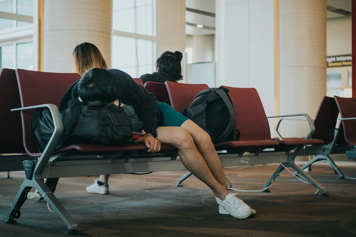 An airport departures waiting area with rows of maroon seats, round columns and tall windows, two travelers asleep on their backpacks