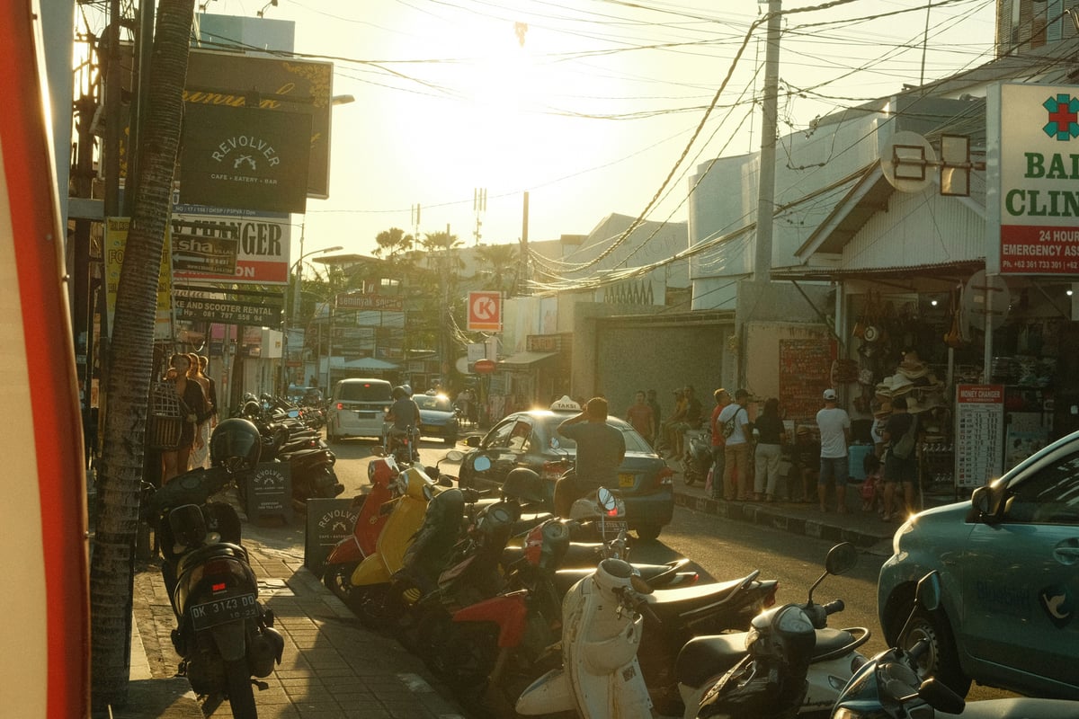 A street in Bali in low evening sun, packed with scooters and cars between shopfronts and tangled overhead cables