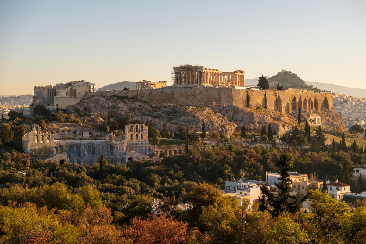 The Acropolis rock with the Parthenon on top, seen from a wooded hill to the southwest in warm afternoon light, with Lycabettus hill behind