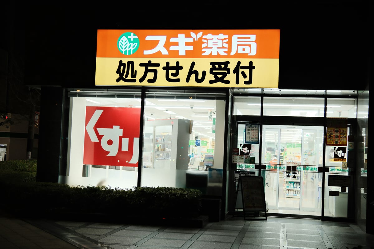 Sugi Yakkyoku drugstore storefront lit up at night, with the illuminated pharmacy sign, a red medicine panel in the window and shelves visible through the glass door