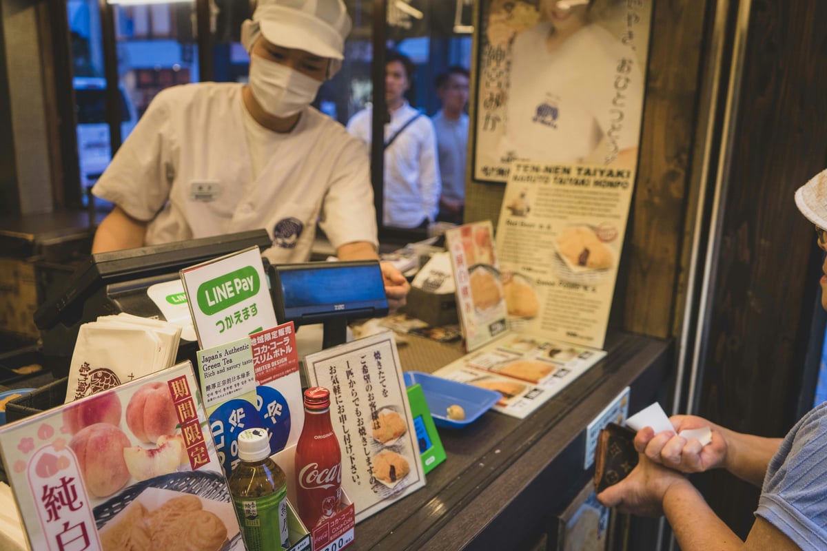 Shoppers browsing the shelves of a brightly lit Japanese drugstore aisle stocked with cosmetics and medicines
