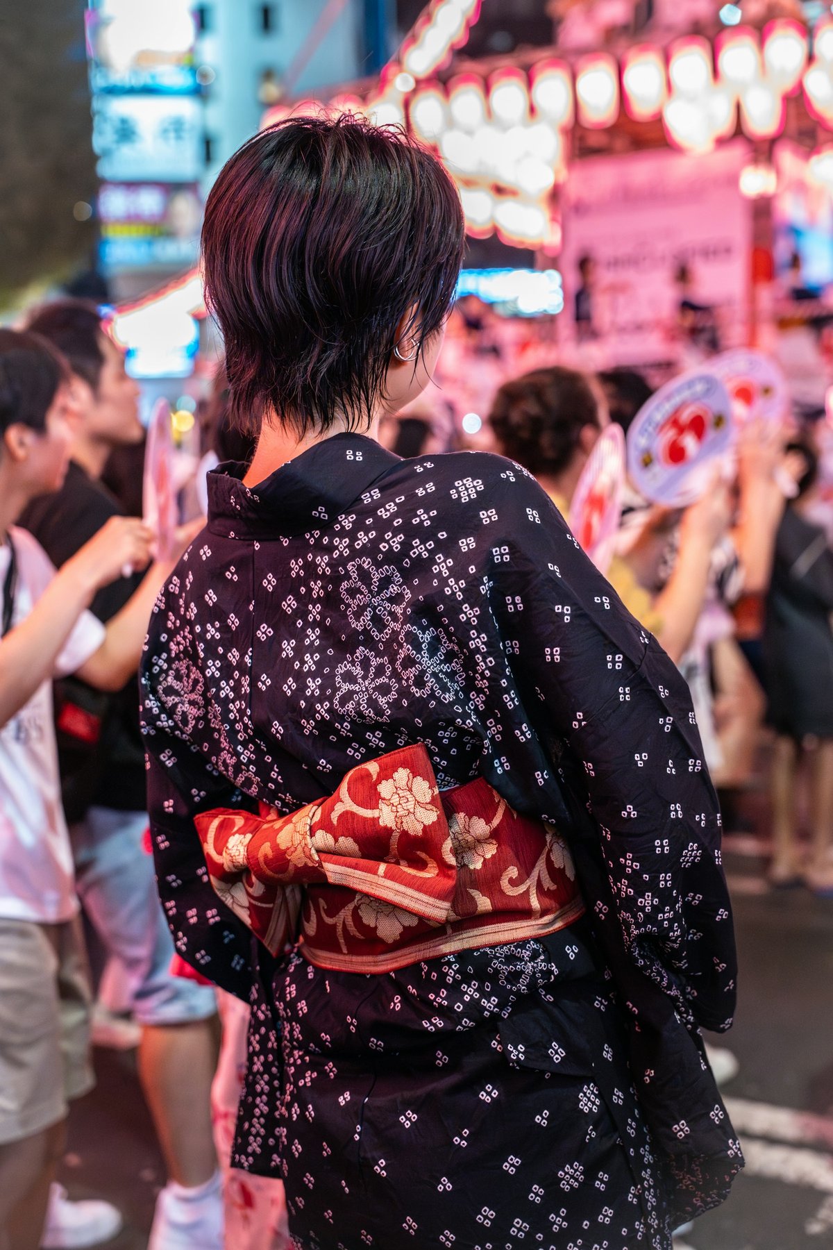 Woman in a patterned yukata, back view, facing a lantern-lit festival crowd