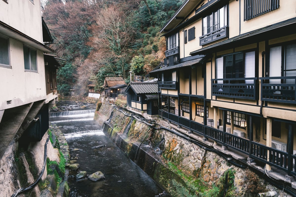 Wooden ryokan buildings stacked above a narrow river gorge in winter, snow on the roofs and steep wooded slopes