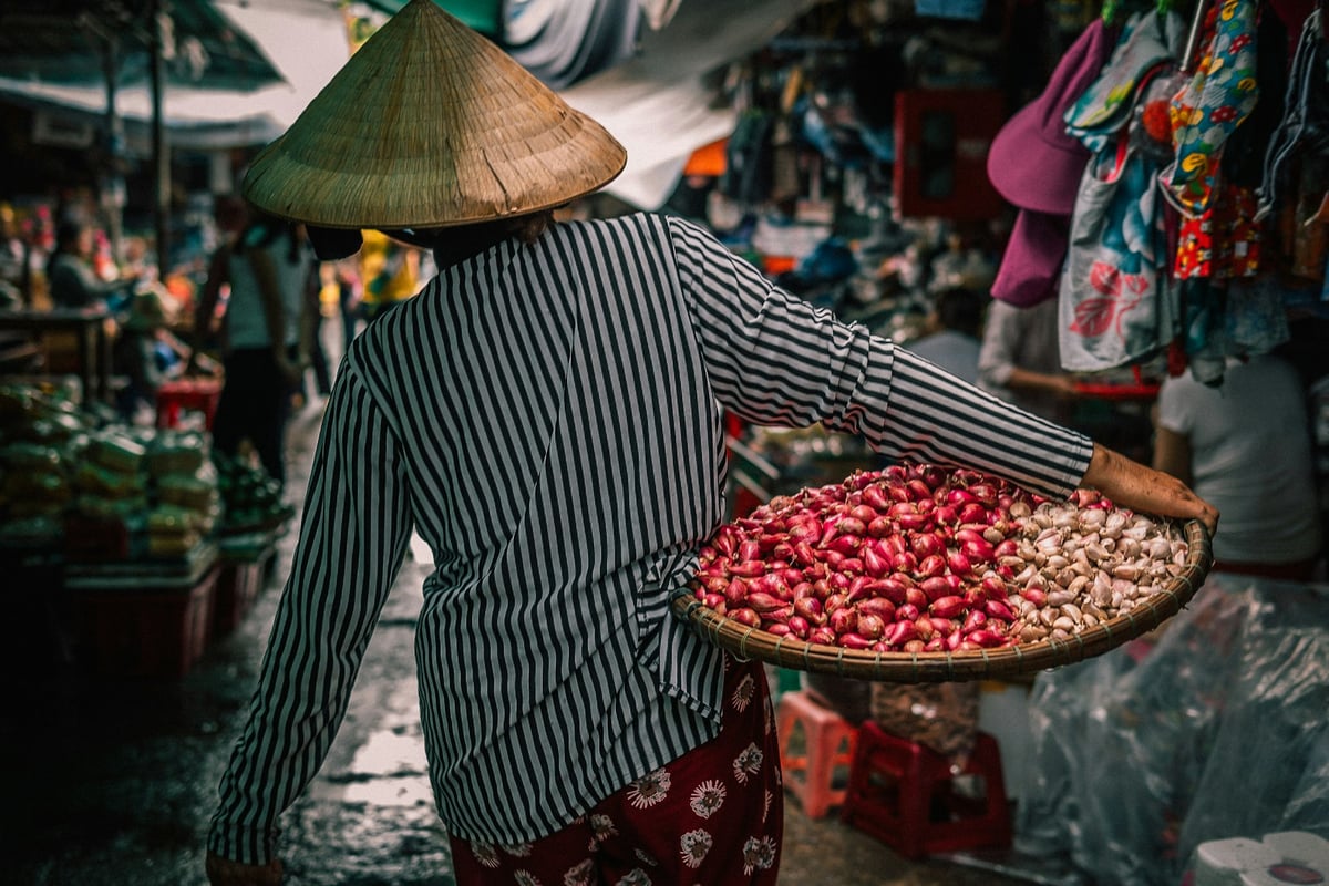 A market vendor in a conical hat carrying baskets of nuts and produce
